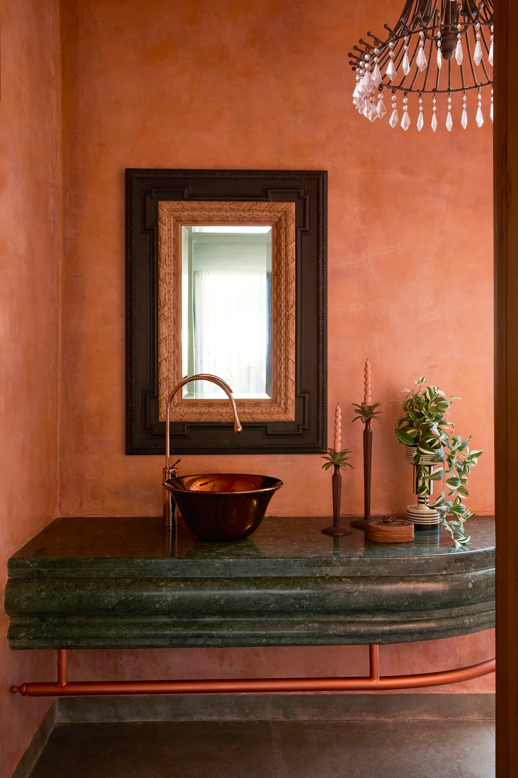 Orange walls surround a dark green floating counter with a copper-toned vessel bowl, layered dark mirror, hanging crystal-like light and small decorative objects.