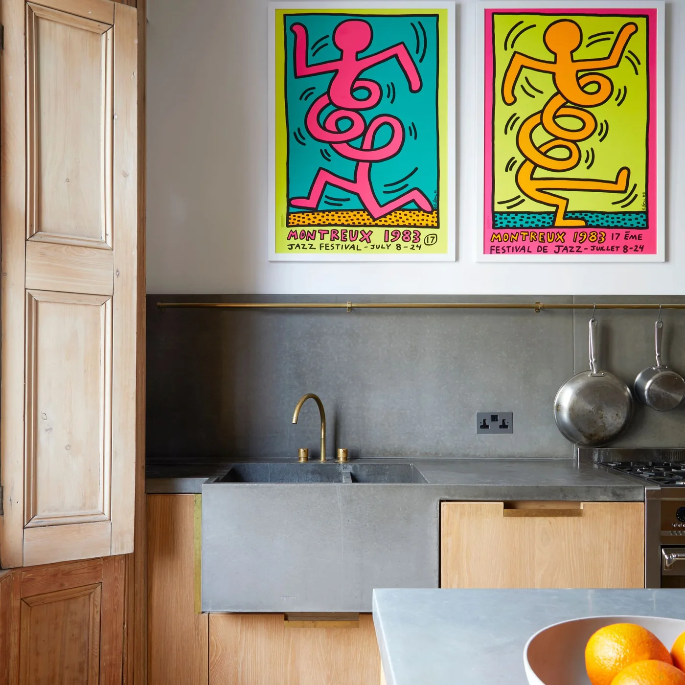 Two colorful framed prints hang over a gray kitchen backsplash beside wooden cabinets and a gold-colored rail