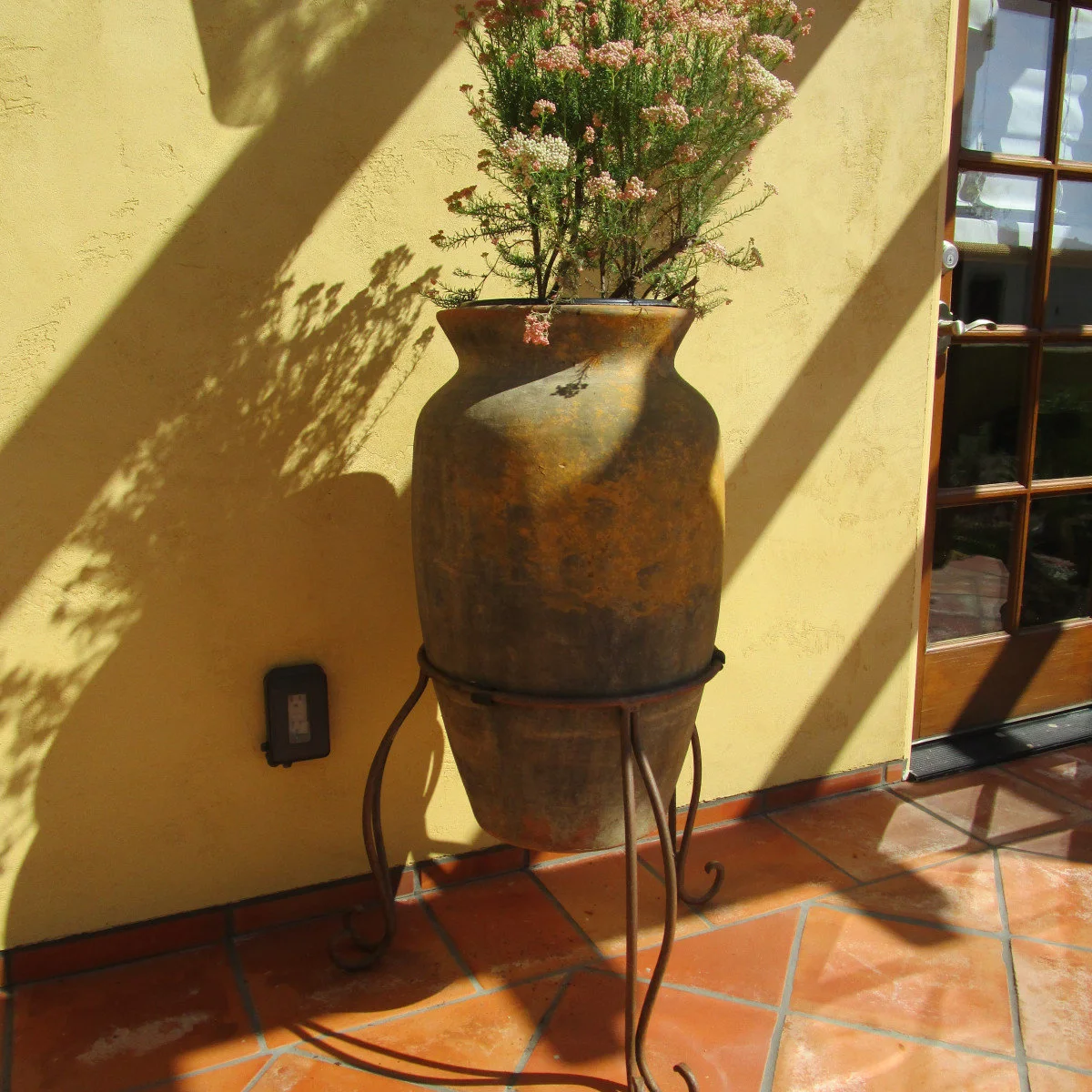 Large warm-toned pot holding small pink flowers on a curved metal stand against a yellow wall beside a wood door.