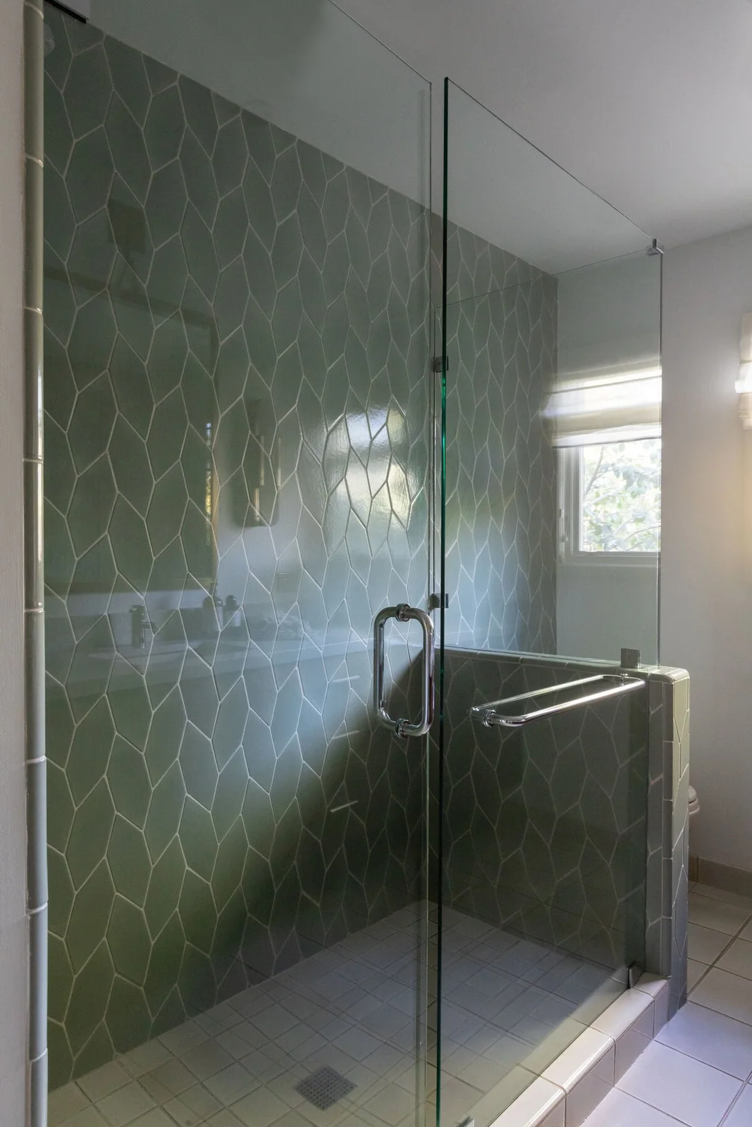 Sage picket tiles form a braided pattern inside a glass shower with a half-height wall, a window and pale floor tiles.
