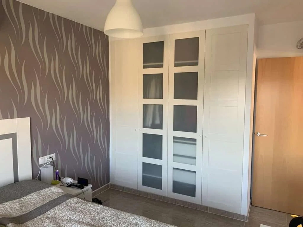 White fitted PAX wardrobe with glazed central doors and solid outer doors beside a patterned bedroom wall