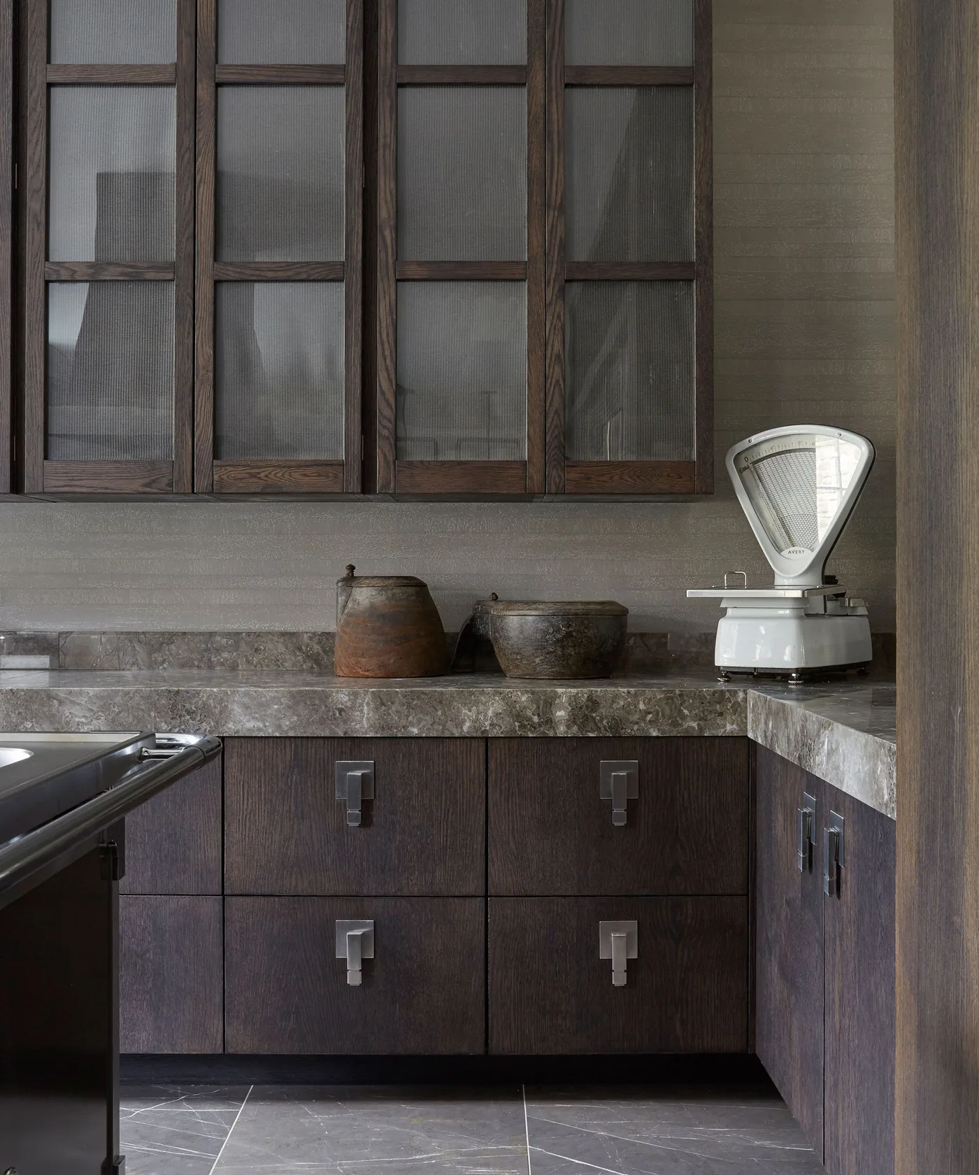 Dark brown kitchen with textured glazed upper doors, square metal handles, gray-brown veined counter and backsplash, and pale weighing scale.