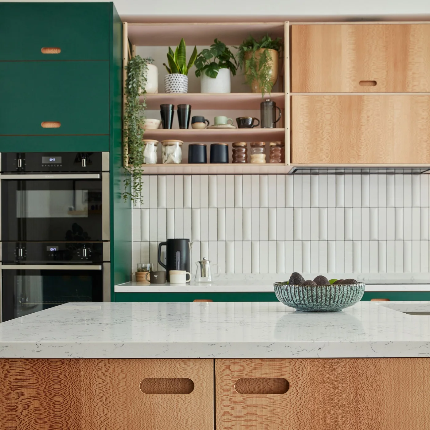 White rectangular tiles stand vertically beneath wooden shelves beside green kitchen cabinets and a pale counter