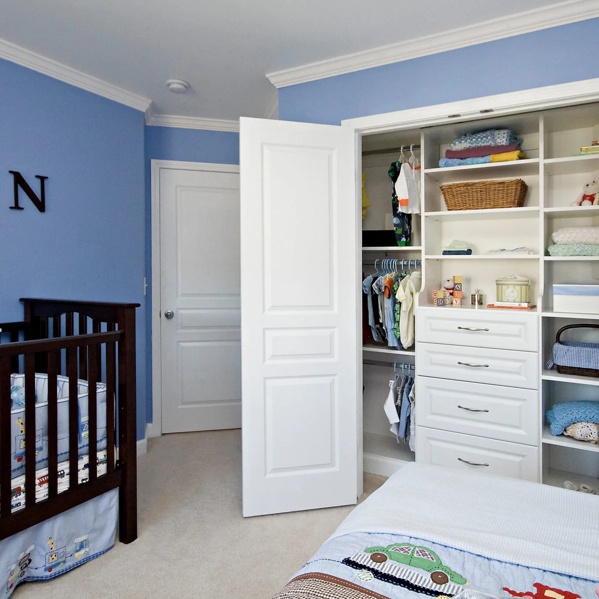 Blue nursery with an open white closet containing drawers, hanging rails and shelves beside a dark crib