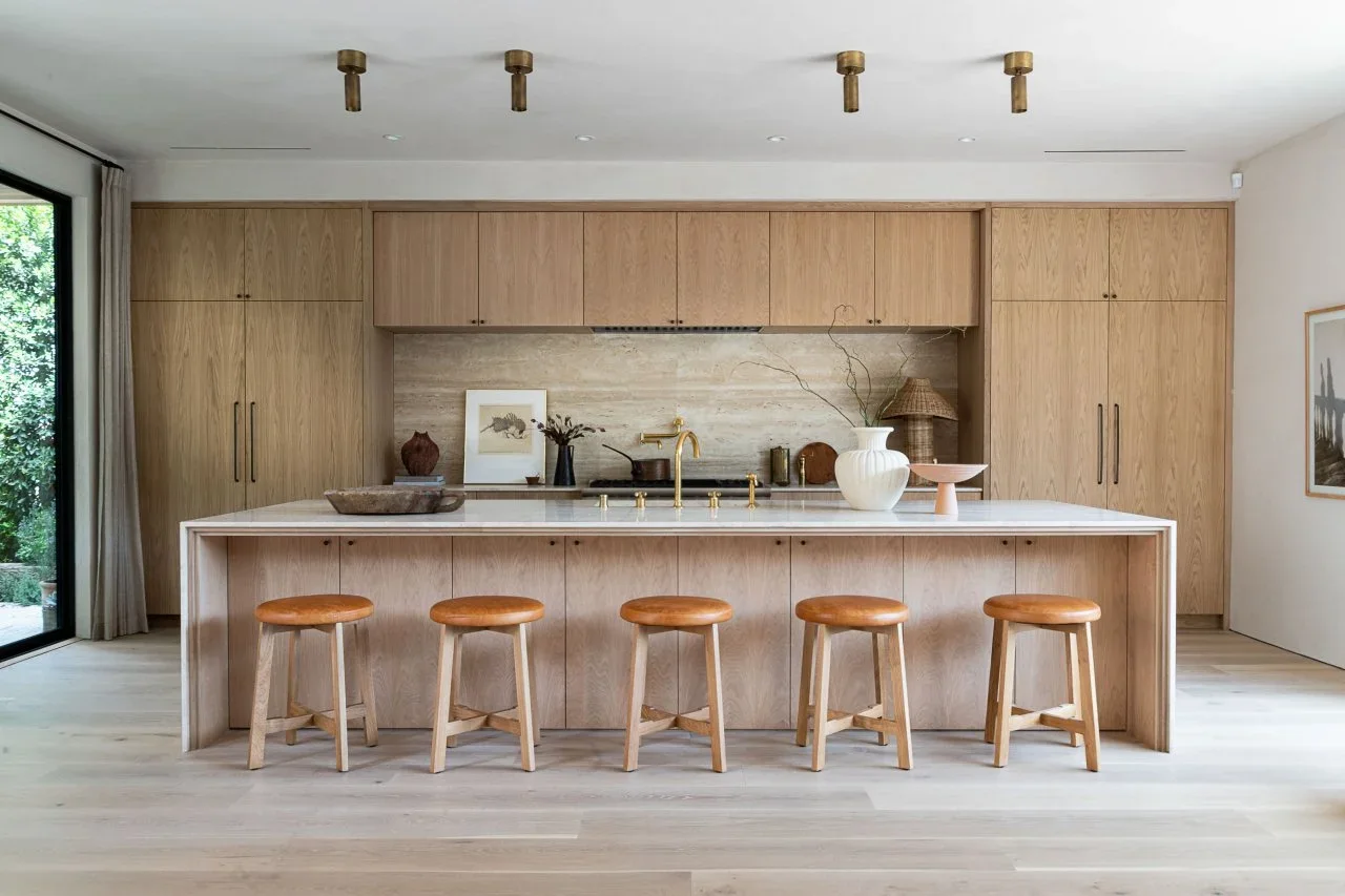 Flat white oak cabinets cover a kitchen wall behind a long island with five backless stools and a veined backsplash.
