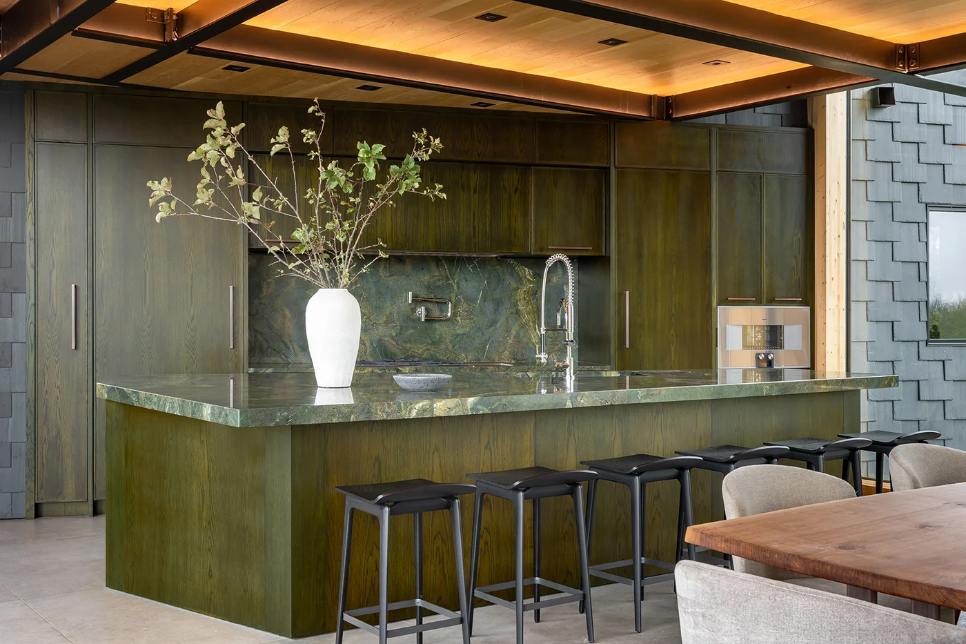 Dark olive cabinets with visible grain surround a green-topped island with dark stools beneath a warm wooden ceiling