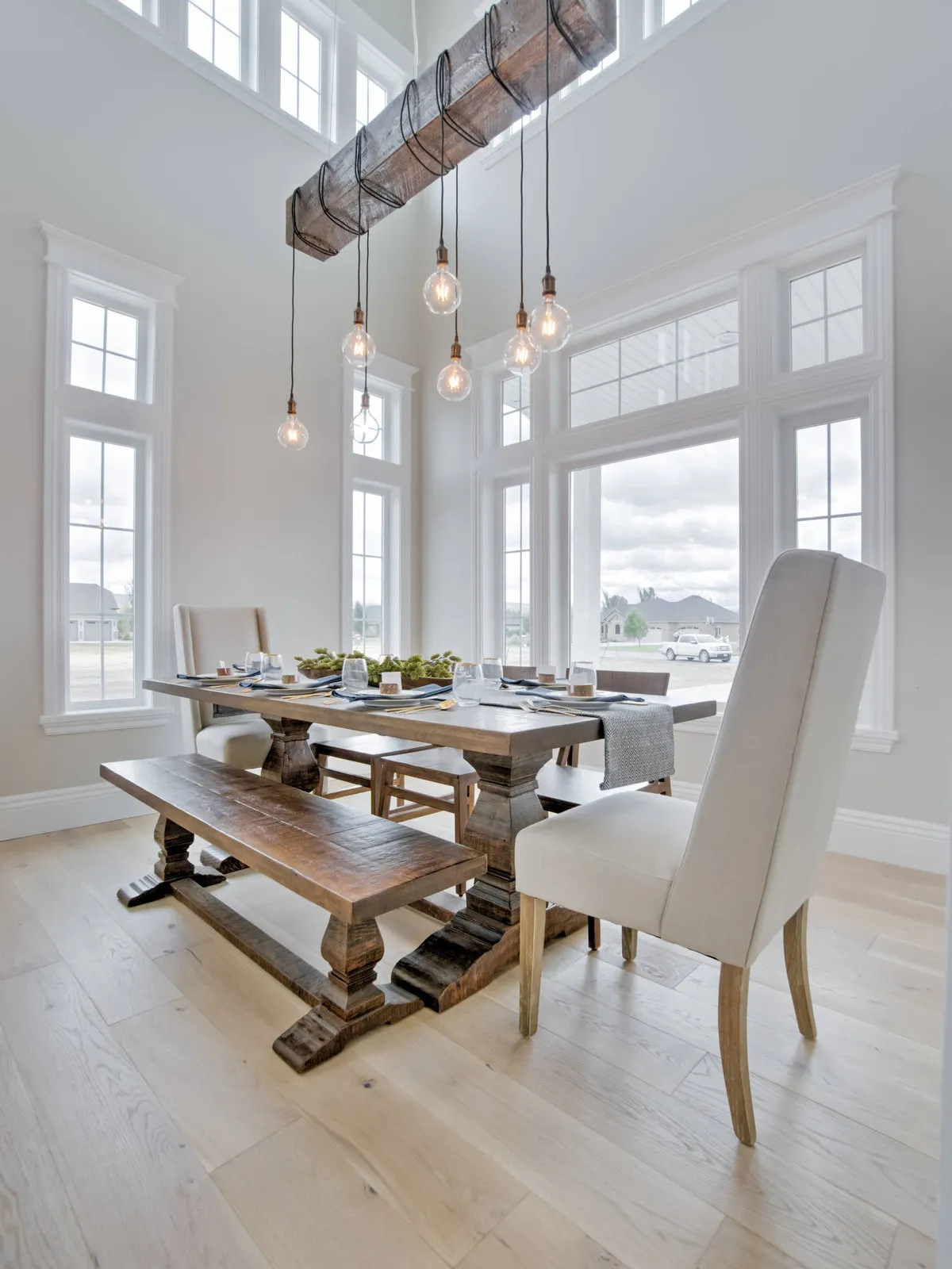 A broad timber table stands beneath hanging bulbs beside pale upholstered chairs.
