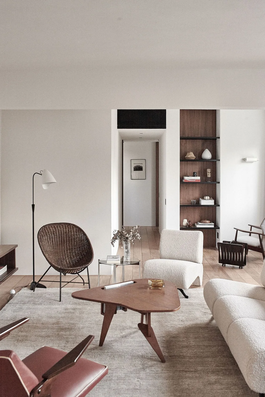 A pale living room has a curved sofa, wood coffee table, open woven chair and smaller upholstered seats beside a narrow wood shelf