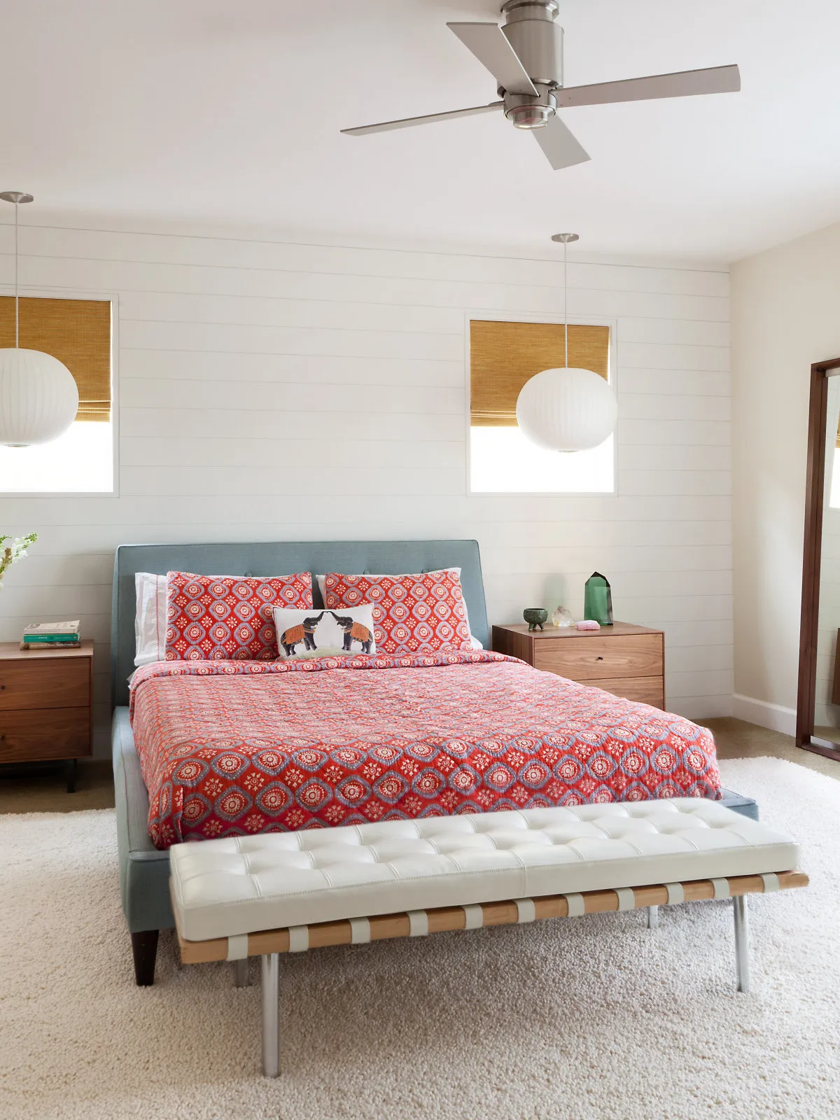 A red patterned quilt covers an upholstered bed beneath pale walls and a ceiling fan.