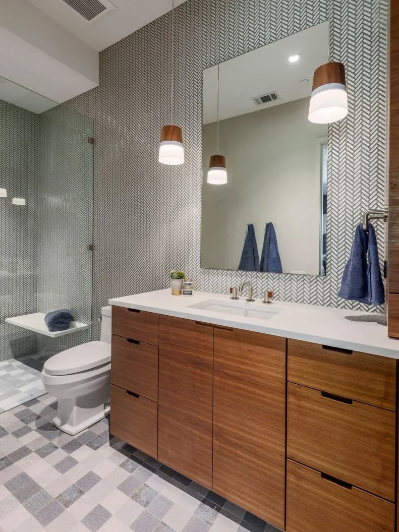 Flat wood bathroom cupboards and drawers stand below a white sink counter beside fine herringbone-patterned walls, a glazed shower and gray patchwork floor tiles