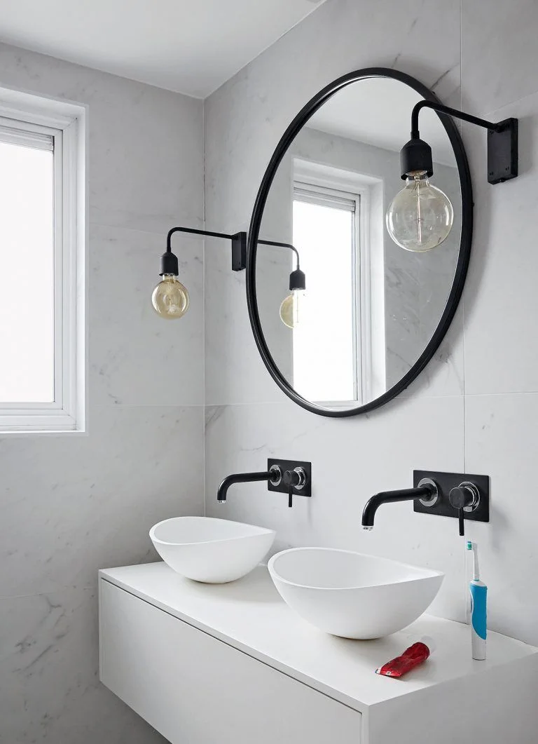Two white bowl basins sit on a white floating vanity below a black round mirror with black wall taps and exposed-bulb lights