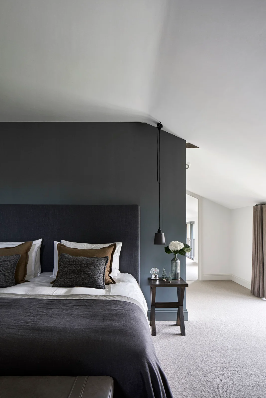 A charcoal headboard wall and gray bedding sit beside a pale sloped ceiling, a small wood bedside table, a hanging dark light and a bright window area.