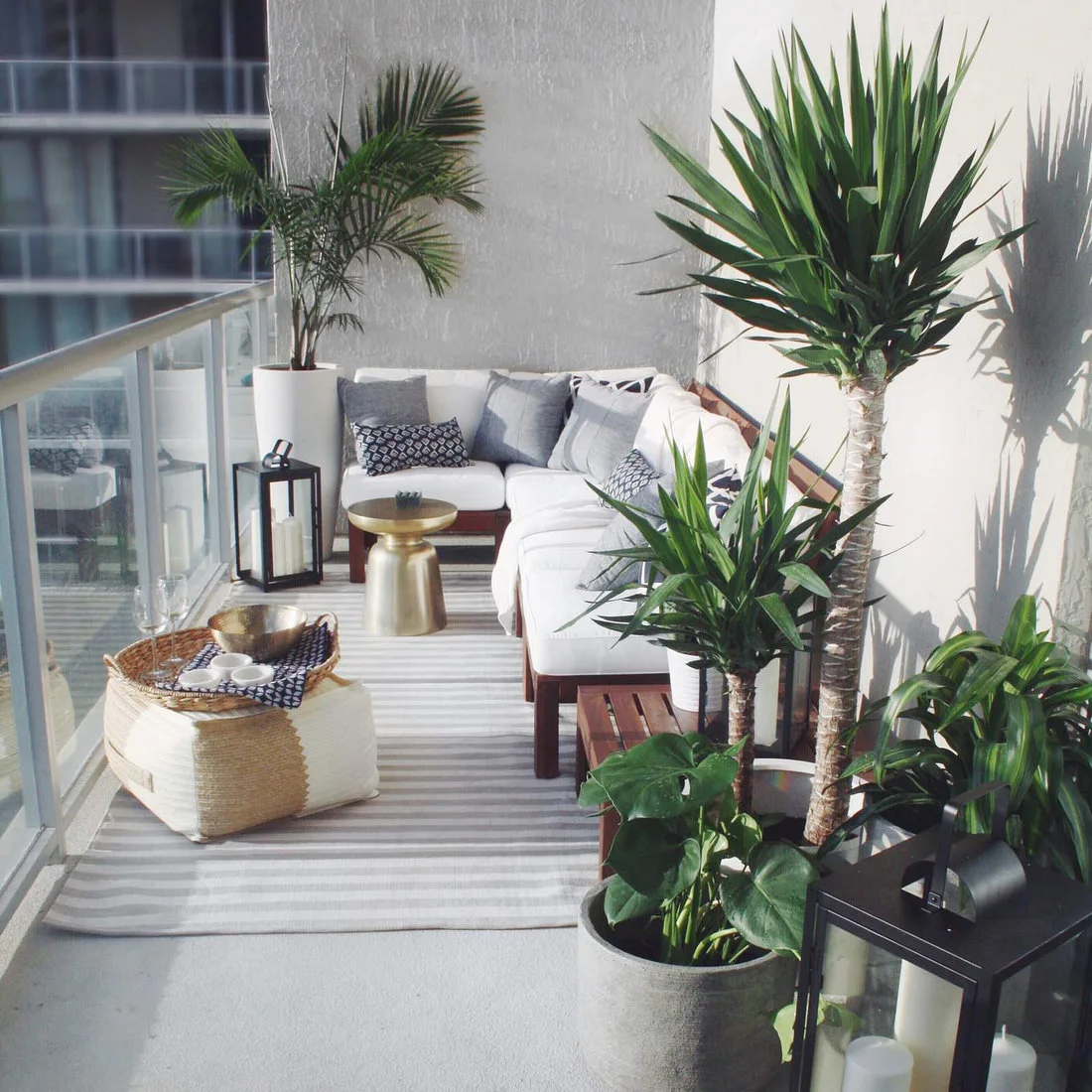 White-cushioned corner seating sits against a balcony wall beside tall plants and small gold-colored tables