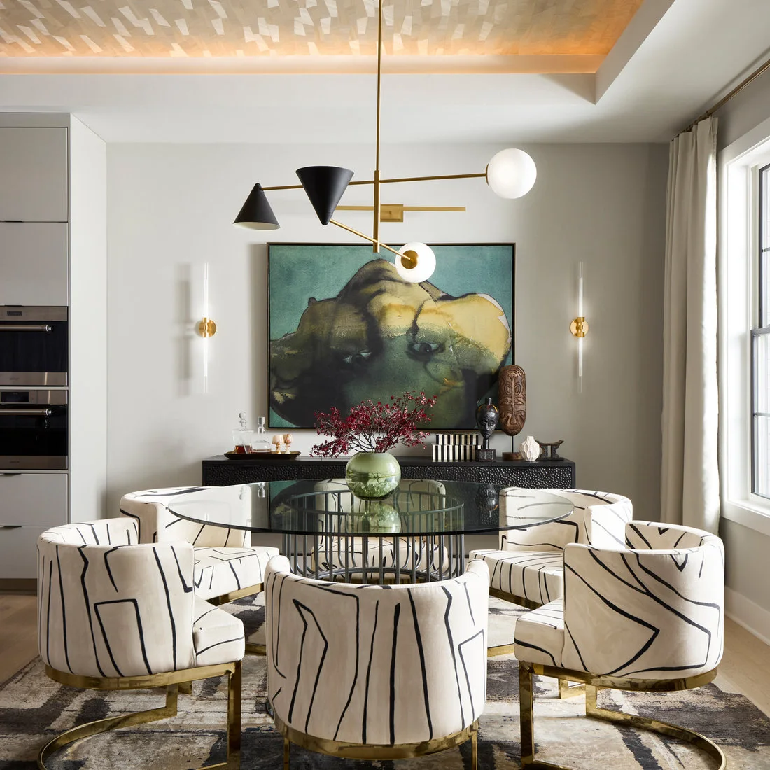A round glass dining table and white upholstered chairs with black line patterns sit beneath a sculptural cone-and-globe light beside green artwork and a dark sideboard