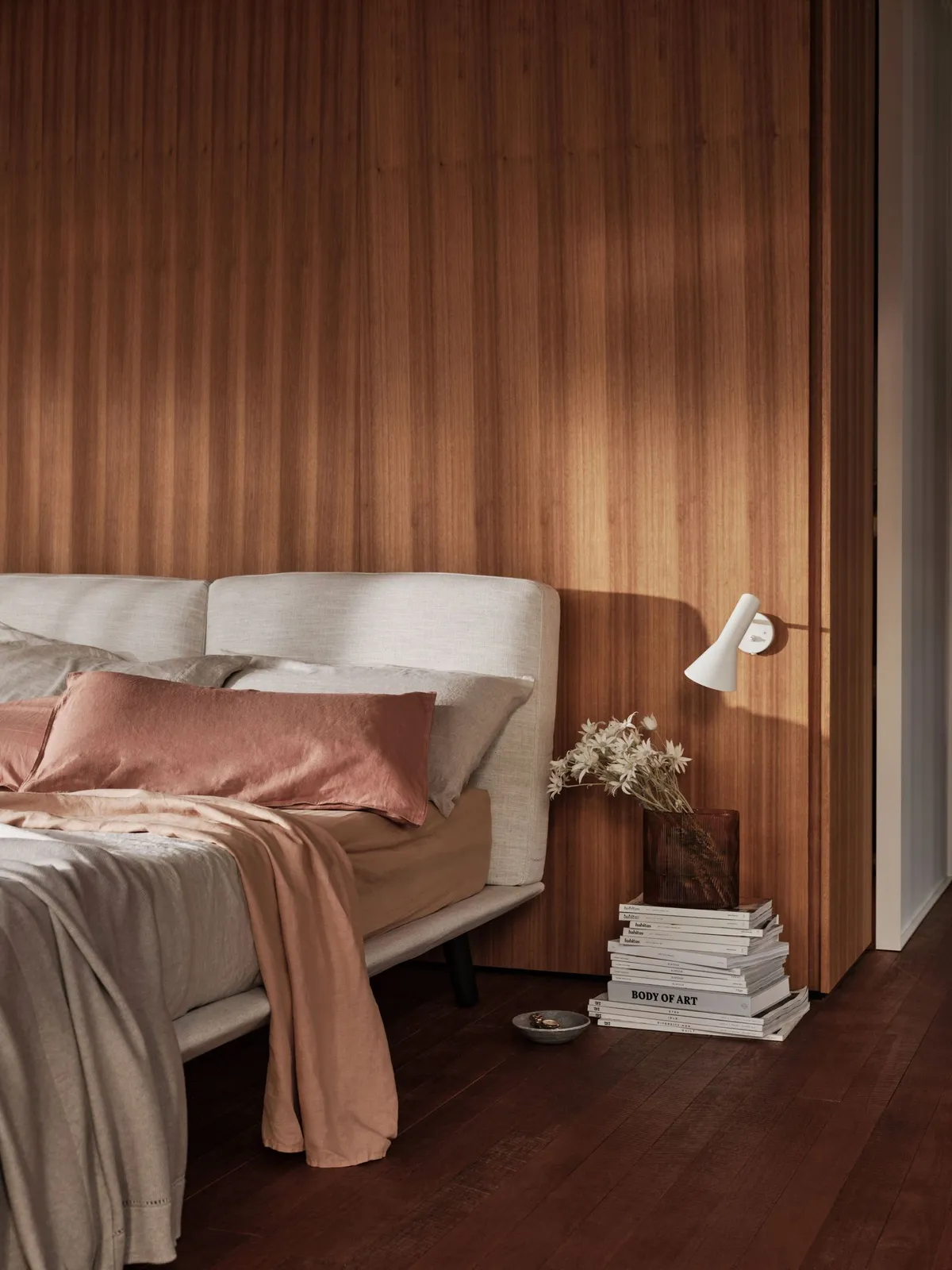 A pale upholstered bed with muted pink bedding stands against warm vertical wood panels beside a small white angled wall light and books stacked on the floor.