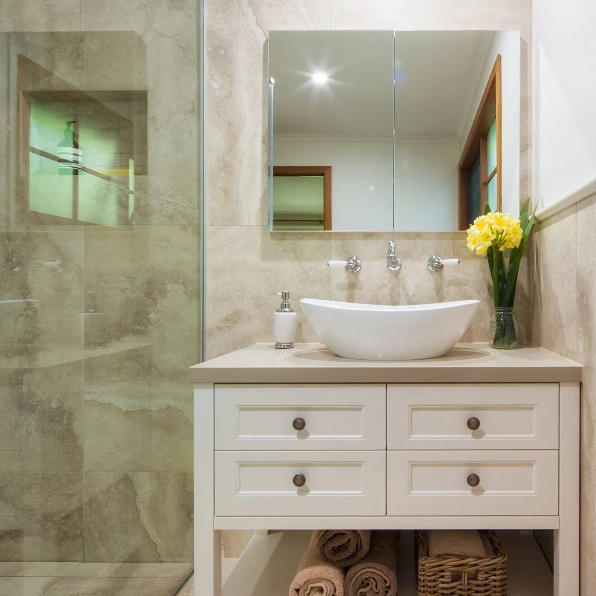 A curved white vessel bowl sits on a small white vanity with drawers and an open towel shelf beside a glass shower screen.