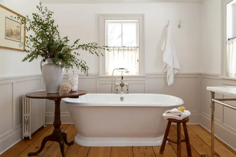 Pale bathroom with freestanding white tub, paneled lower walls, wood floor, dark round table and small stool