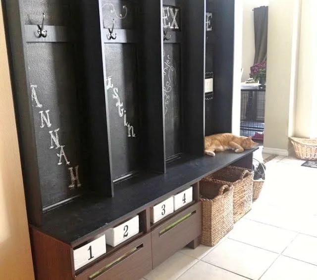 Dark chalkboard-style entryway lockers with names, black hooks, lower numbered bins and woven baskets beside a low storage unit.
