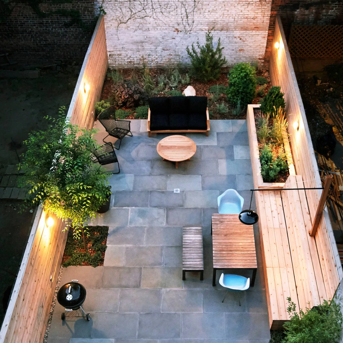 A dark outdoor loveseat and two chairs surround a round wood table at one end of a narrow planted courtyard.