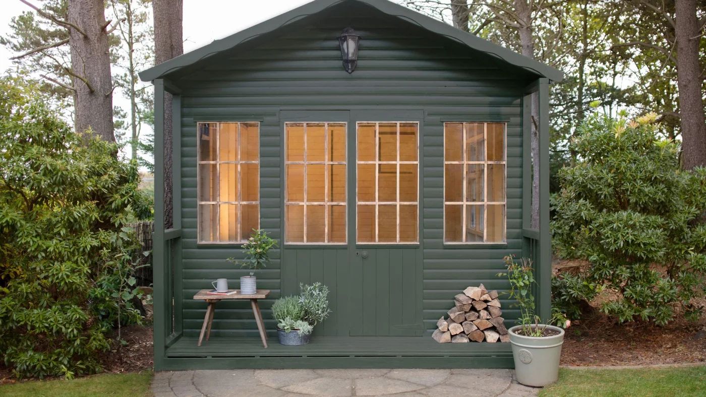 A dark green garden building with glazed doors stands between shrubs with a few pots at its entrance