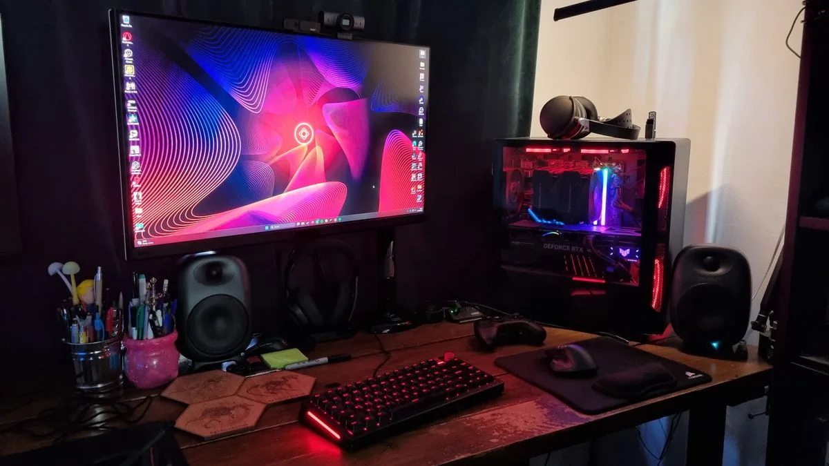 A wood desk with one monitor, a red-lit computer tower, black speakers, a red-backlit keyboard and dark curtains.