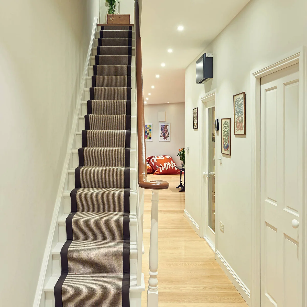 Narrow entrance hallway beside a straight staircase, with framed pictures, pale walls and a striped stair runner.