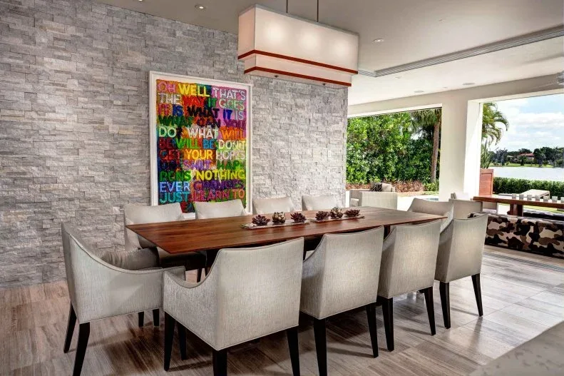 Dining room with a warm wood table, pale upholstered chairs, gray textured wall, colorful text artwork and rectangular pendant