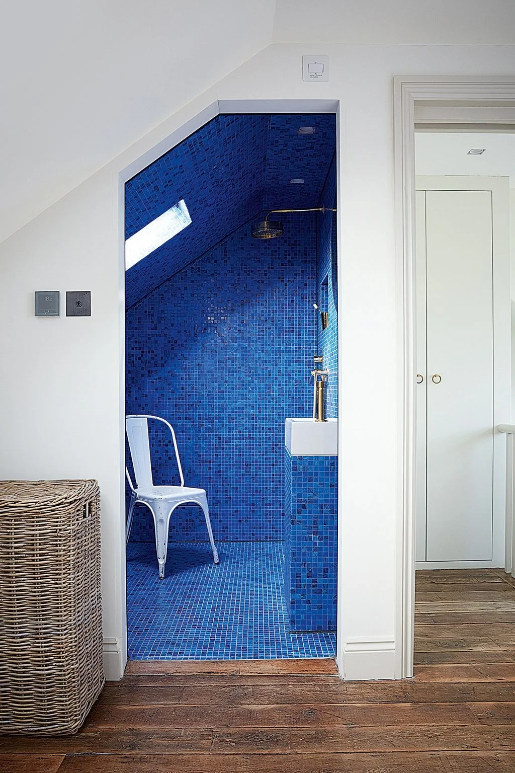 An open attic shower lined in bright blue mosaic tiles has a sloping ceiling, a skylight and brass-colored fittings.