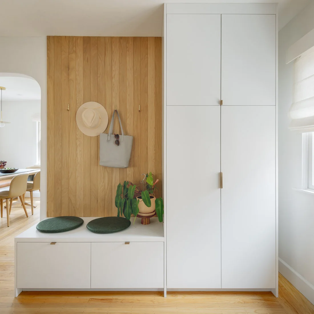 A white bench cabinet with round green seat pads sits beside a tall white cupboard and a pale wood hook wall near a window.