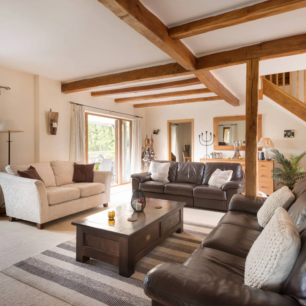 Two dark brown leather sofas and a pale patterned sofa arranged around a cream rug beneath wood beams.