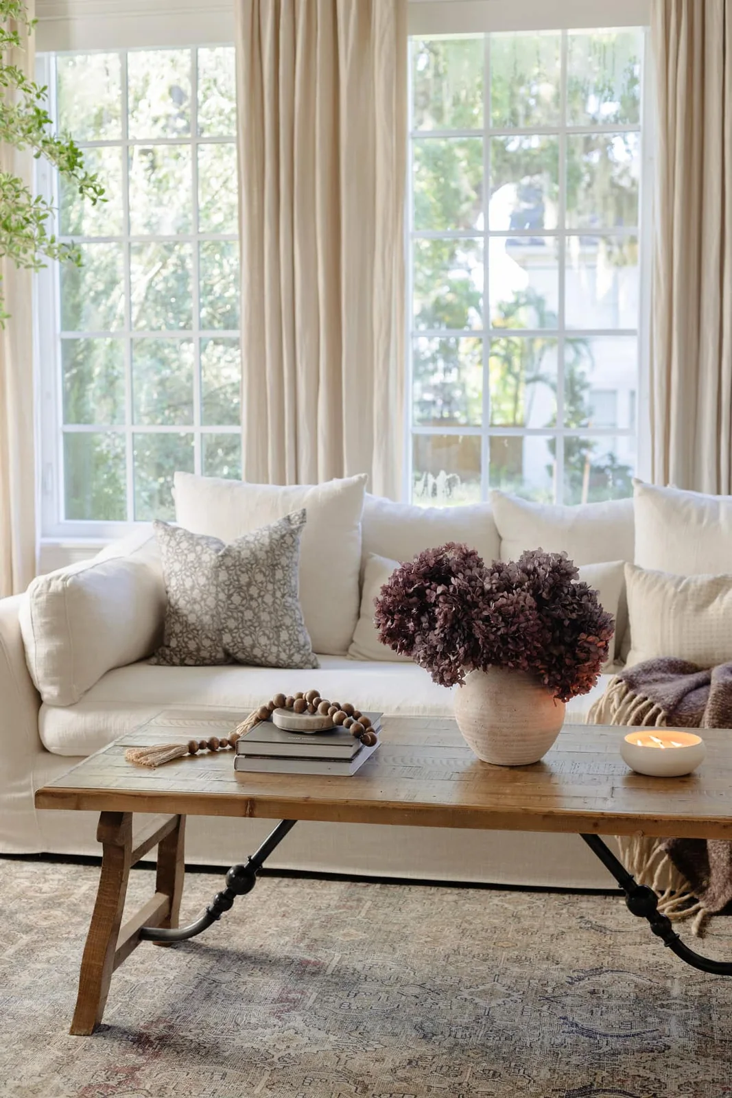 Muted purple hydrangeas fill a rounded pale pot on a timber coffee table in front of a pale sofa and tall windows.
