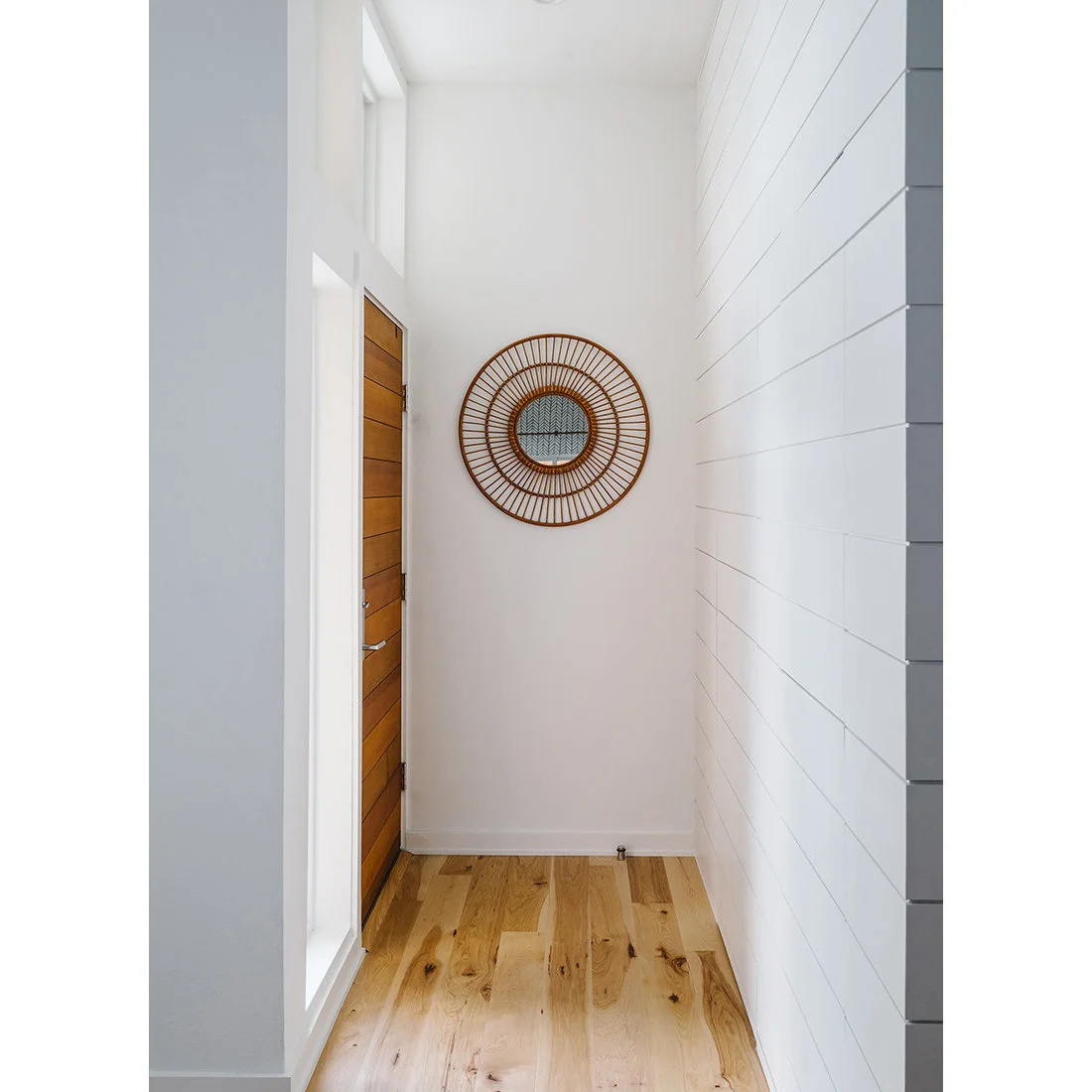 A round woven mirror hangs at the end of a narrow white passage beside a wood door and pale wood floor.
