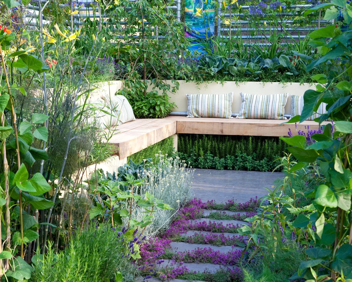 A timber corner bench with striped cushions sits among deep planted beds and upright growing supports