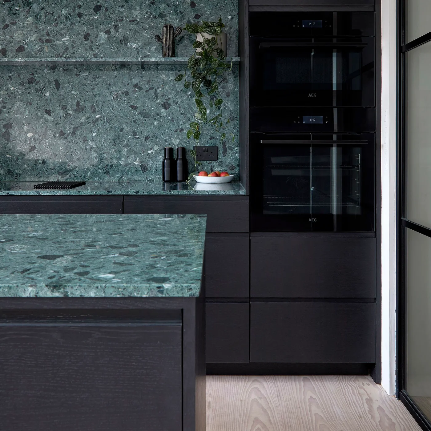 Black kitchen cabinetry surrounds a green patterned backsplash and countertop beside stacked black ovens