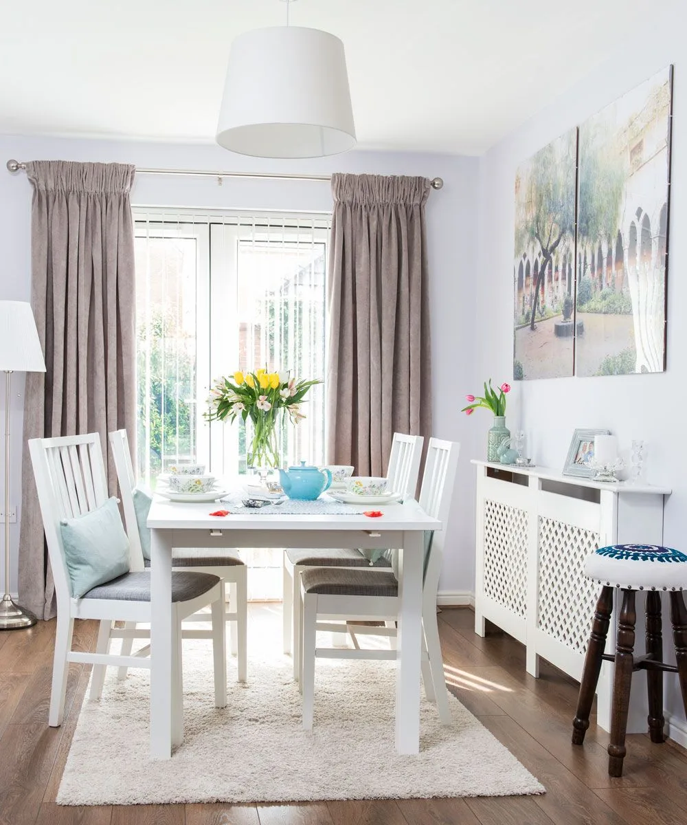 Taupe gathered curtains beside glazed doors, with a white dining table and chairs, wooden flooring, a pale pendant shade, and colorful wall artwork.