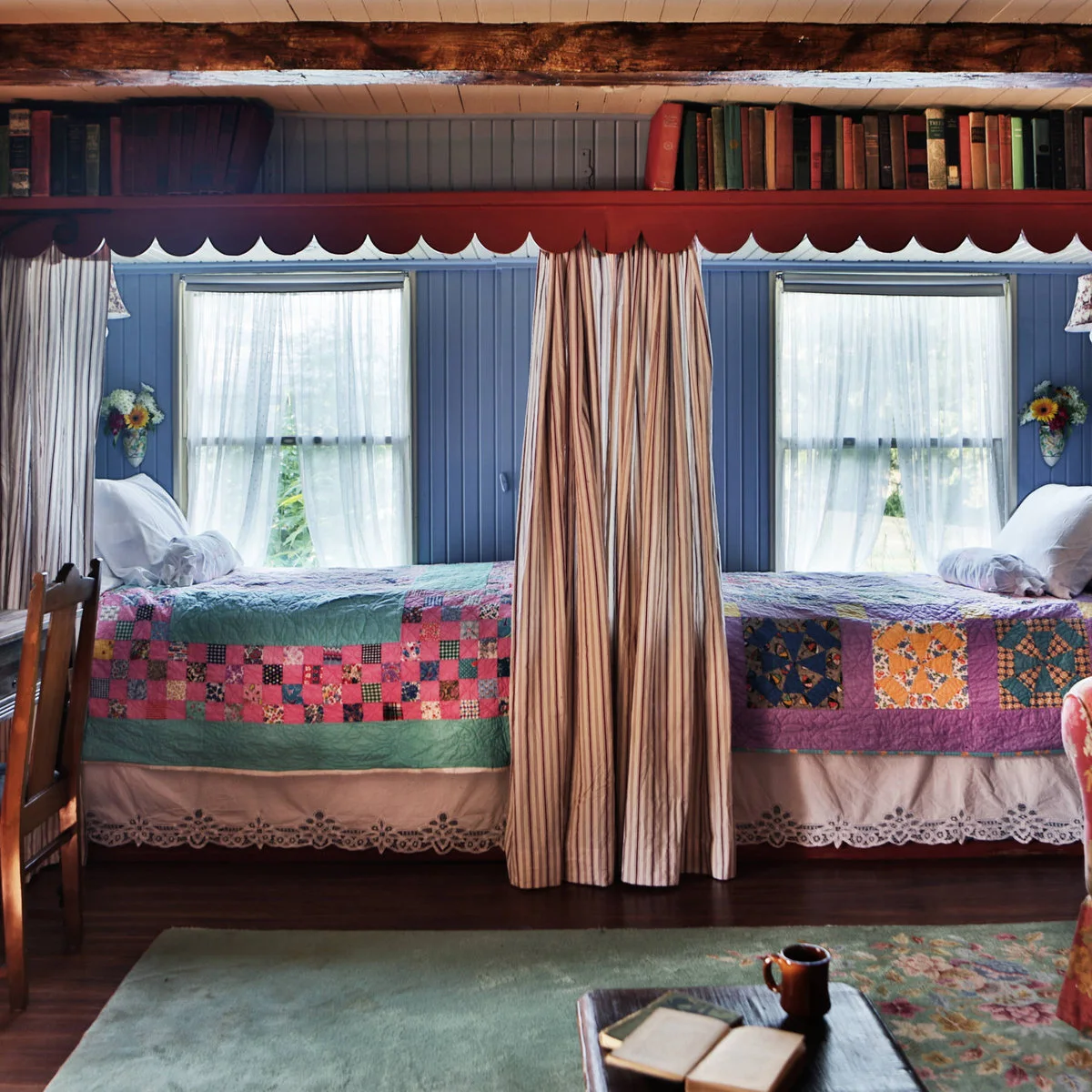 Two end-to-end beds beneath windows with patchwork quilts, blue paneling and a central curtain