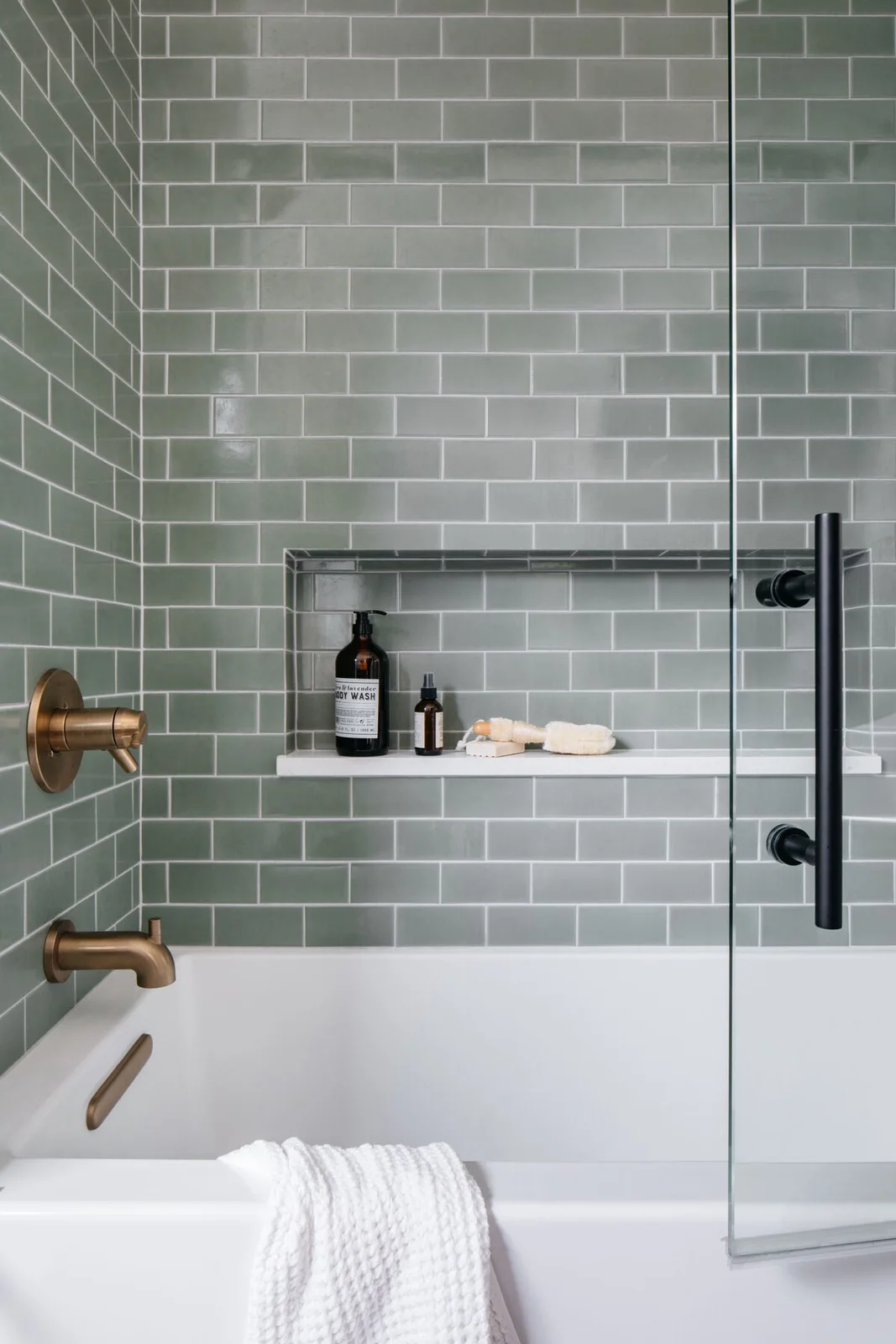 A white bathtub has a gray-sage subway-tile surround with a broad recessed niche, brass fixtures and a clear glass screen.