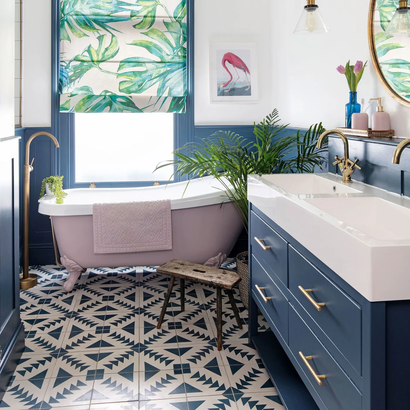 A deep blue vanity and blue paneled walls sit beside a pink clawfoot bath and patterned floor