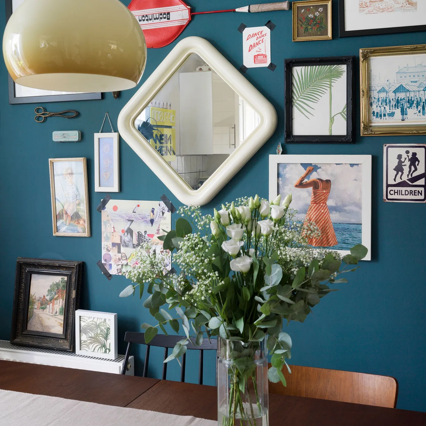 Pale rounded diamond-shaped mirror among framed artwork on a teal wall behind a wood dining table