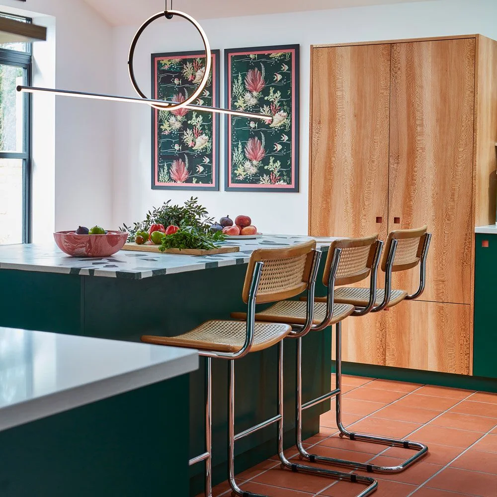 Two tall dark botanical prints beside a wood-front kitchen cupboard and green island with woven stools.