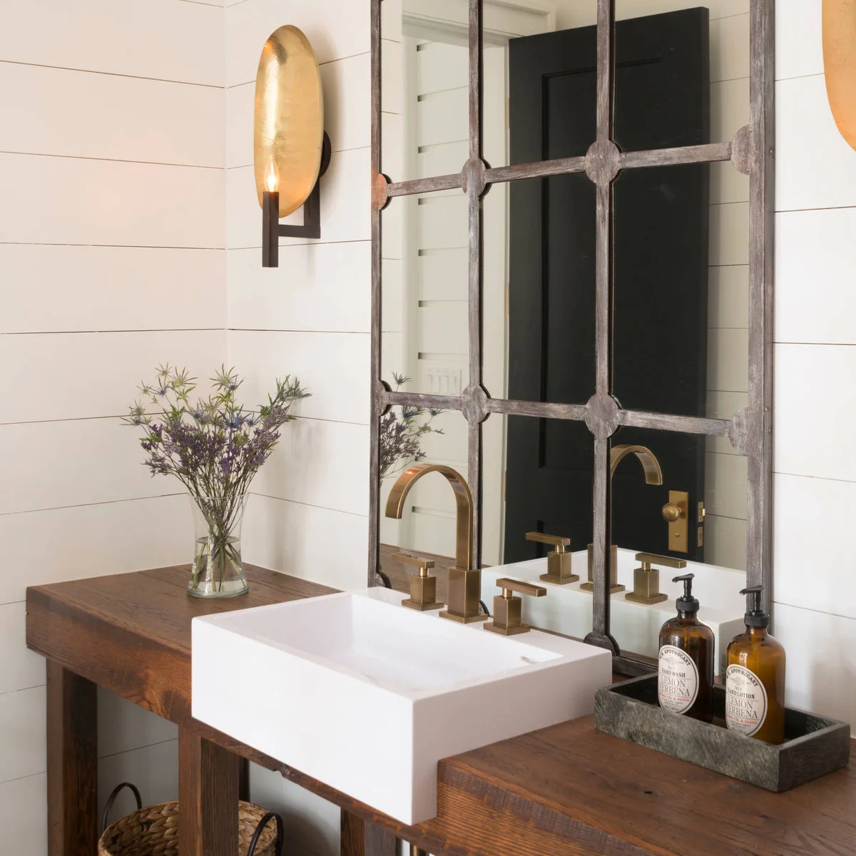 Wood vanity with a white rectangular basin, two amber pump bottles on a dark tray and a small vase of purple flowers.
