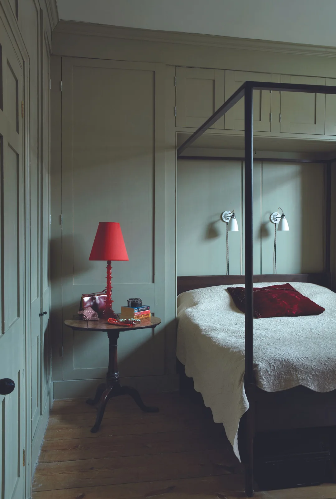 Gray-sage wall paneling surrounds a dark four-poster bed beside a red lamp and a pale bedcover.