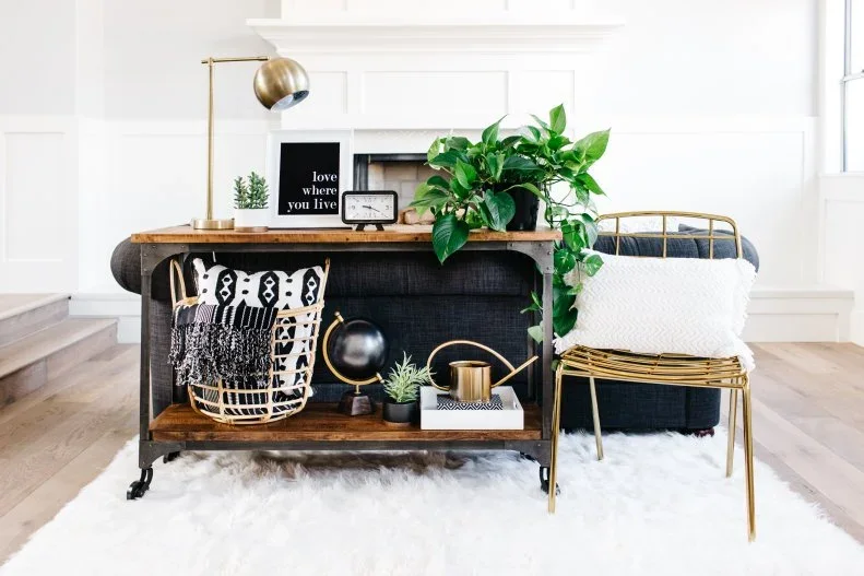 A sofa console holds a gold-toned desk lamp, trailing greenery and a basket of black-and-white cushions.