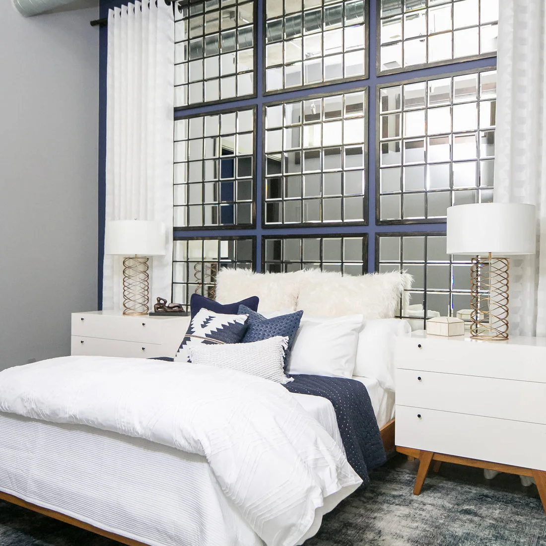 White bedding and drawer cabinets sit beneath a tall dark window grid framed by pale curtains and navy cushions