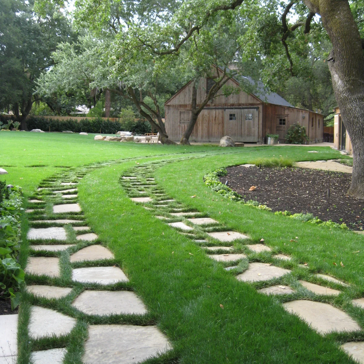 Curving driveway tracks made from pale individual slabs with grass between them leading toward a timber outbuilding.