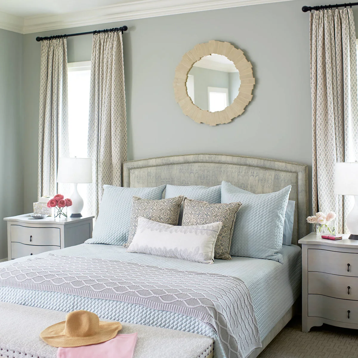 Bedroom with pale blue quilted bedding, a patterned upholstered headboard, cream curtains and a round mirror