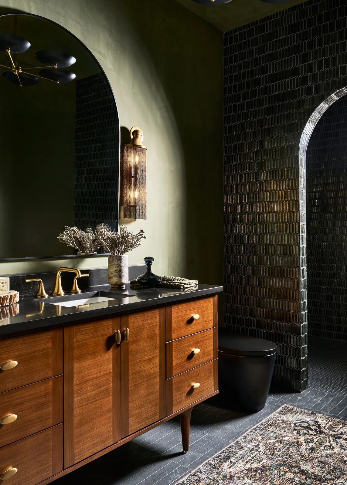 Deep olive walls surround an arched mirror above a wood vanity with a dark counter, beside black fixtures and a small tiled wall section.