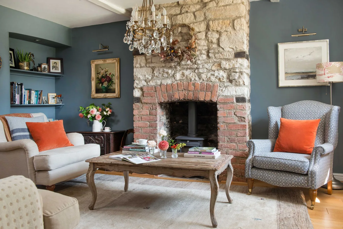 Pale stone chimney breast with dark fireplace, narrow wooden ledge, blue-gray walls and pale seating with orange cushions.