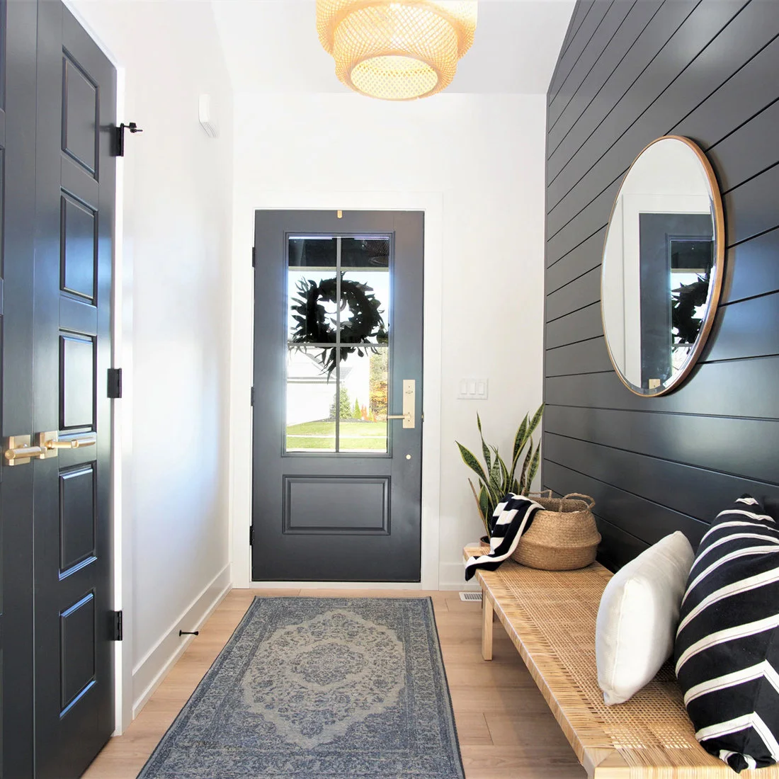 A woven wood bench sits against a dark horizontal-board wall below a round mirror beside white walls, a dark door and a patterned runner.