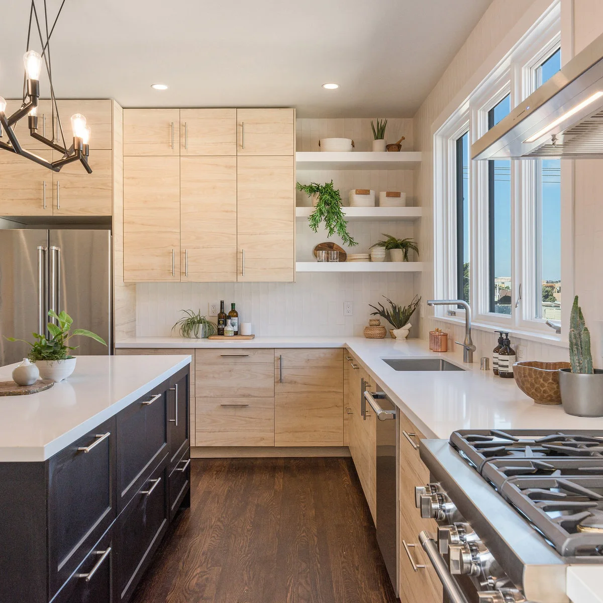 A kitchen sink beneath wide windows with a slim overhead light, pale wood cabinets and open corner shelves.